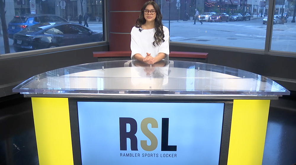 A sports anchor sits at a bright studio desk with large windows behind them, presenting a segment for Rambler Sports Locker.