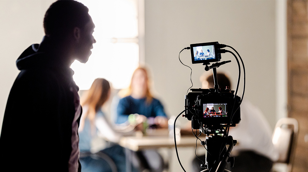 A camera on a tripod films a group seated at a table, while a person stands nearby observing the recording setup.