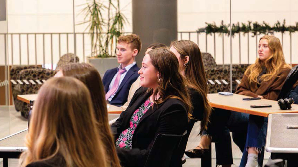 People seated in a classroom-style setting listen attentively during a presentation, gathered around tables in a bright, modern space.