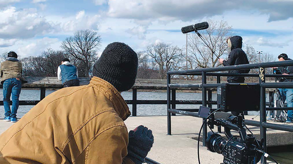  A film crew works outdoors near a waterfront, operating a camera and boom microphone while others lean on the railing in the background.