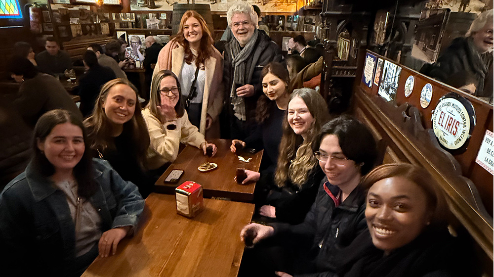 People sit and stand around a wooden pub table, socializing and enjoying drinks in a warm, crowded tavern environment.