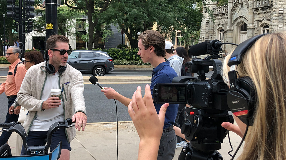 A reporter interviews a man holding coffee as a camera operator films them on a busy sidewalk near buildings.