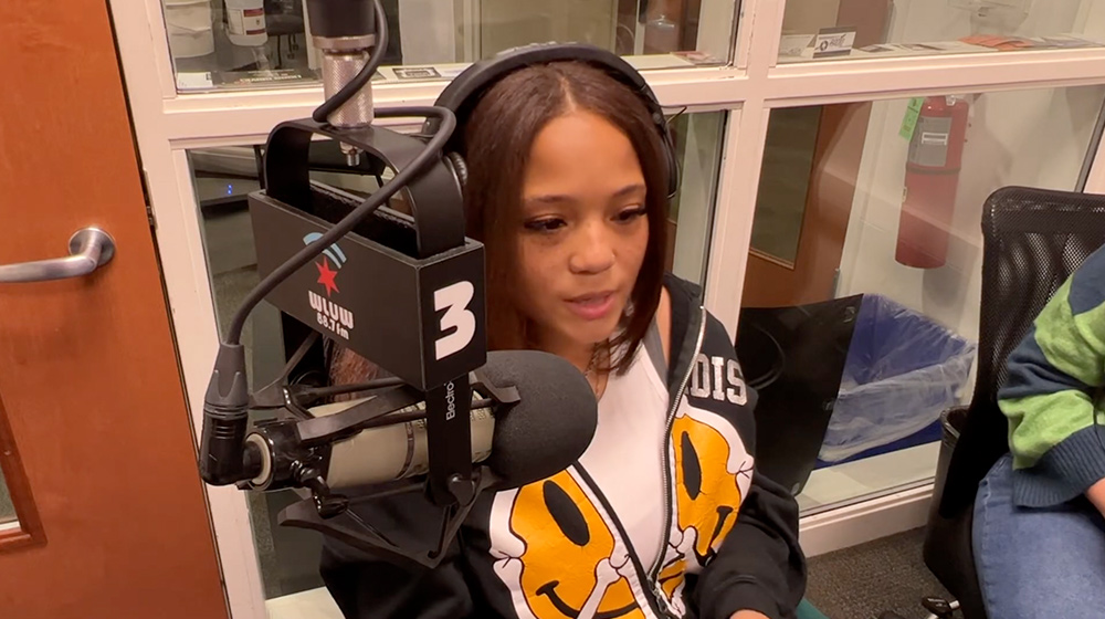 A person speaks into a professional radio microphone inside a studio, wearing headphones while participating in a live or recorded segment.