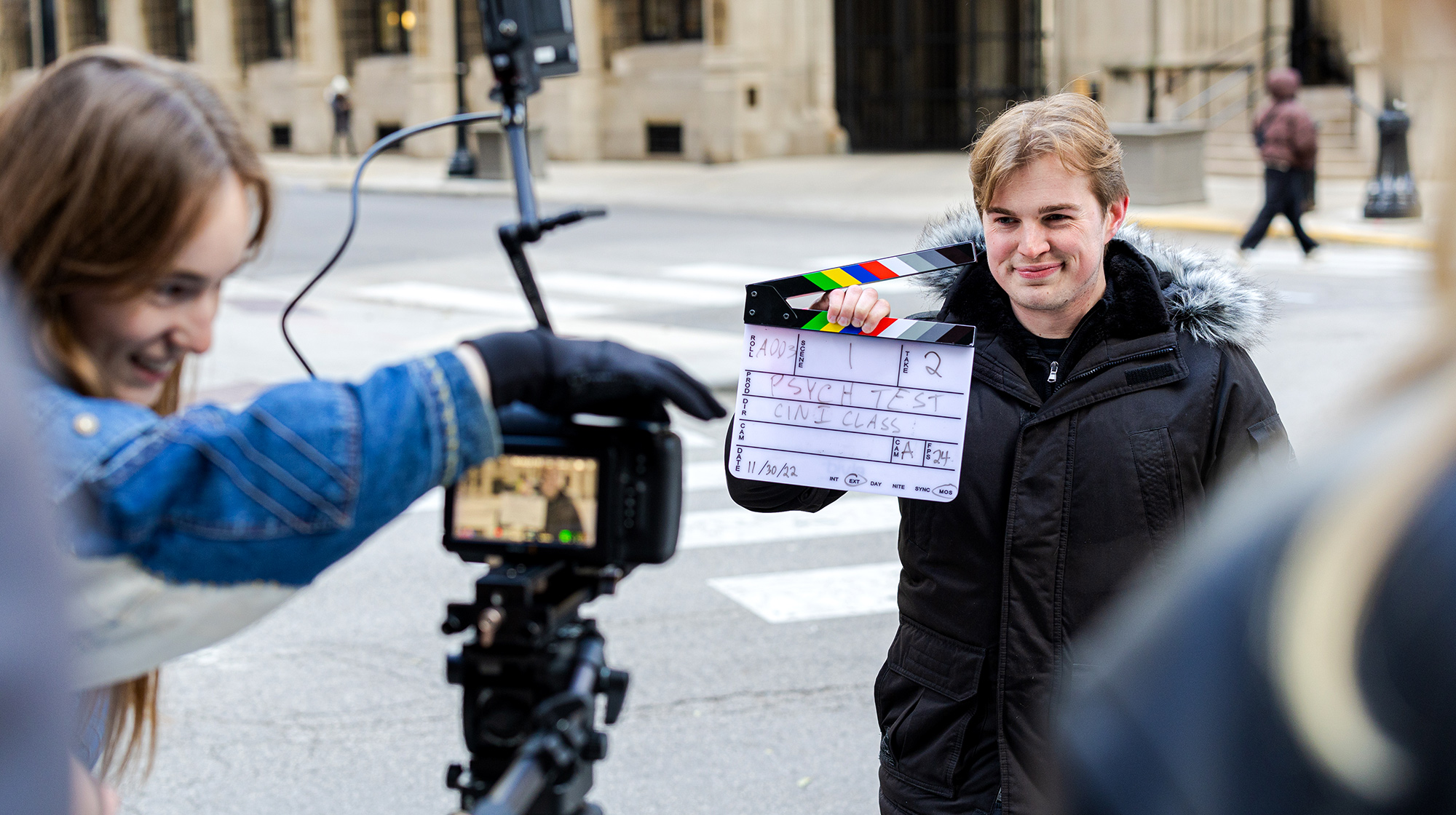 Students film a scene on a city street as one person holds a clapperboard and another records with a professional camera.