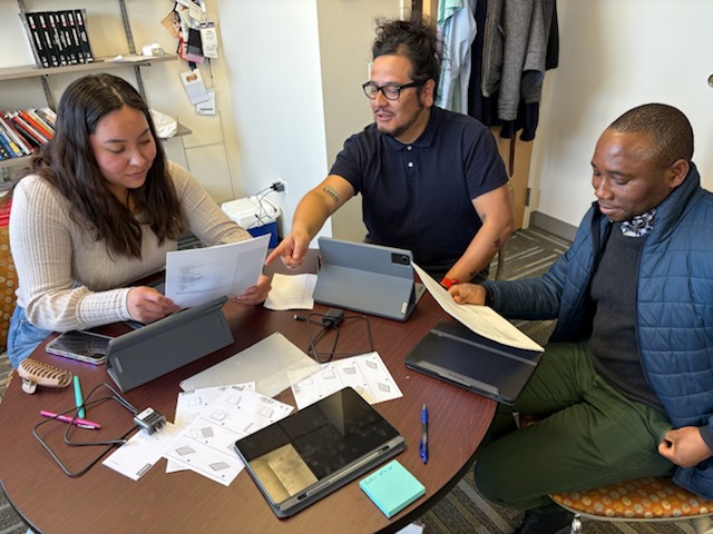 Three people, one woman and two men, working at a desk with paper and computer tablets 