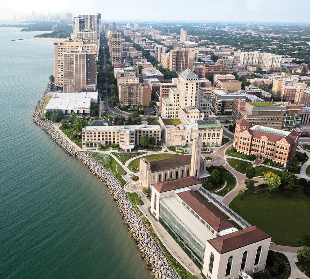 Loyola University Chicago's lake shore campus from the northeast high over Lake Michigan. The lake is an emerald green and the shy is hazy. The campus buildings sit next to lush green grassy spaces nestled against the rocky shoreline of the lake.