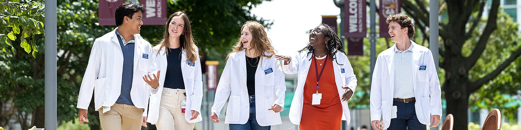 Five medical students, dressed in white lab coats, laugh together as they walk under a canopy of trees and a clear sky.