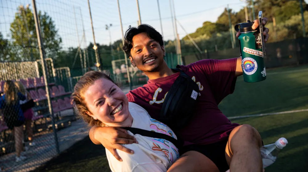 A student carrying another student, both dressed for a soccer match and holding water bottles.
