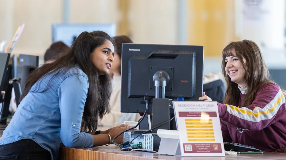 Two women speaking in a library, looking at a computer monitor. 