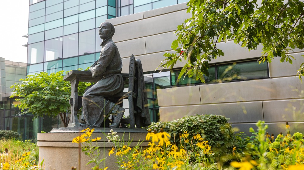A statue of St. Ignatius writing at a desk elevated on a platform among flowers on campus.