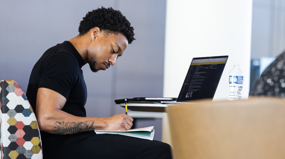A student writes in a notebook and works at their laptop in the library. A student writes in a notebook and works at their laptop in the library.