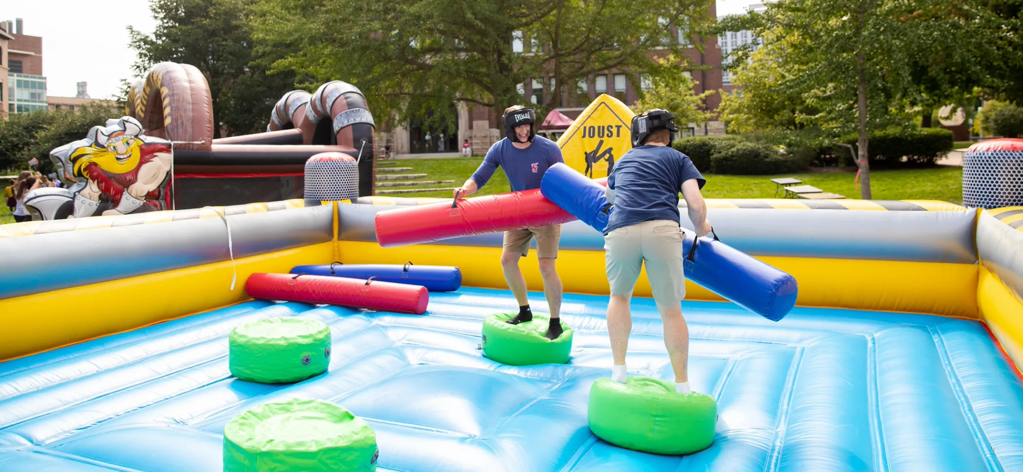 Two people wearing helmets balance on pedestals in an inflatable jousting arena, each holding a padded joust stick, outdoors on a sunny day.