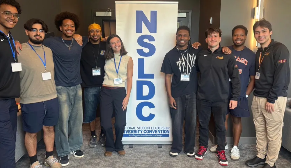 A group of nine people stand smiling in front of a banner for the National Student Leadership Diversity Convention.