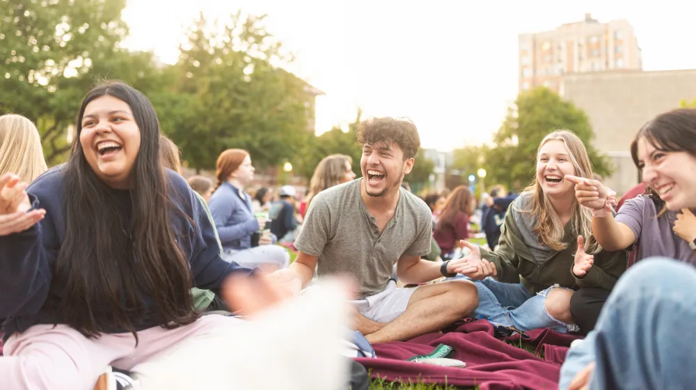 A group of students sitting on a blanket and laughing together outside.