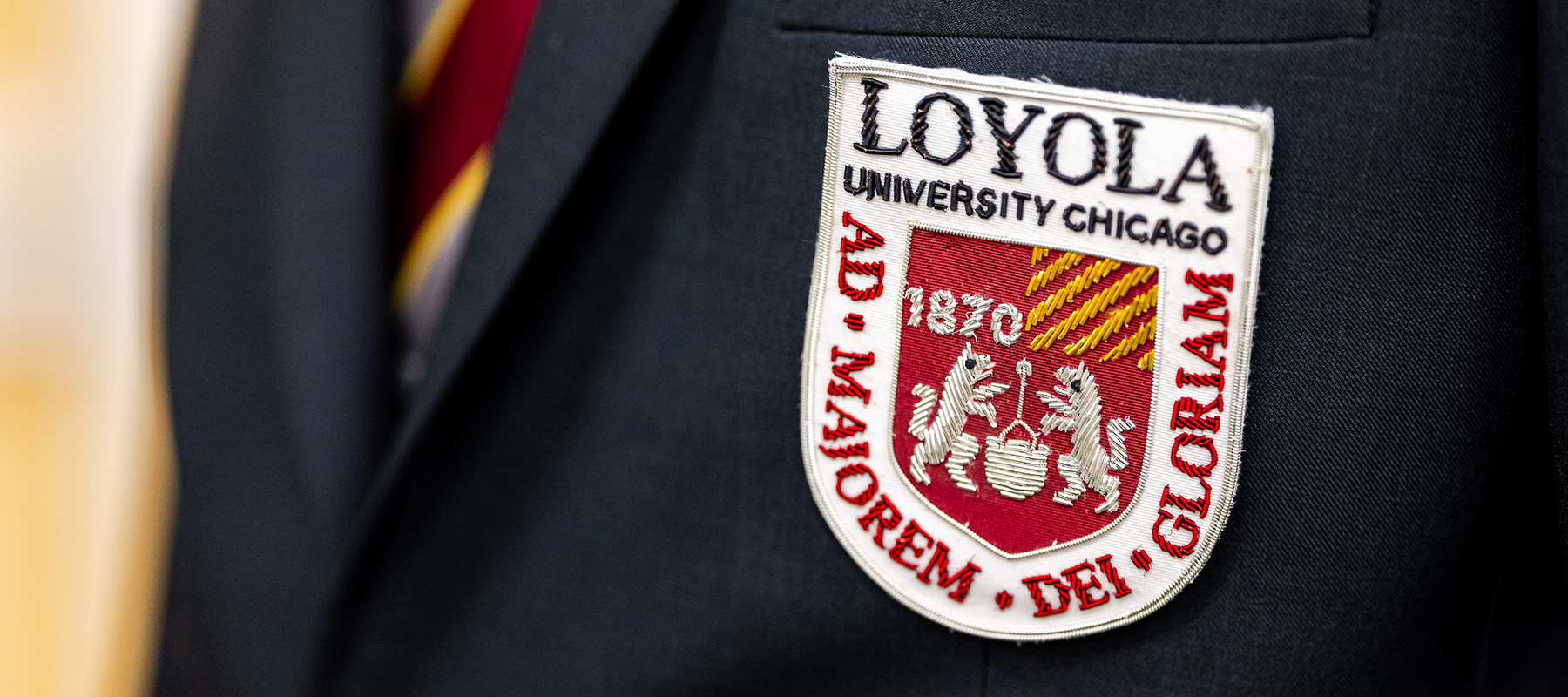 A close up of a sportcoat breast and lapel with an embroidered patch of Loyola's logo and a maroon and gold striped tie.