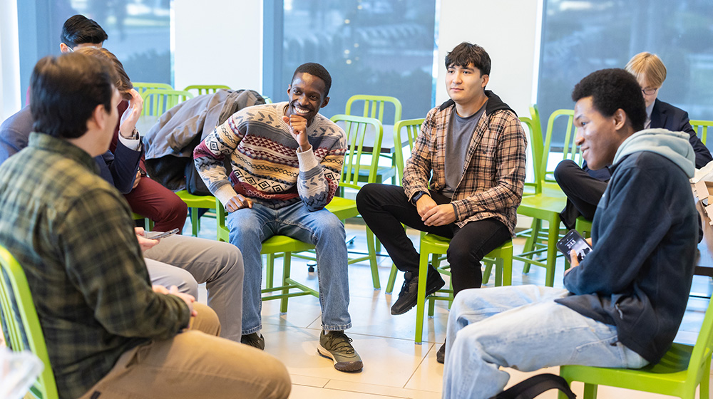 A group of young men talk and smile while they sit on green chairs in a circle.