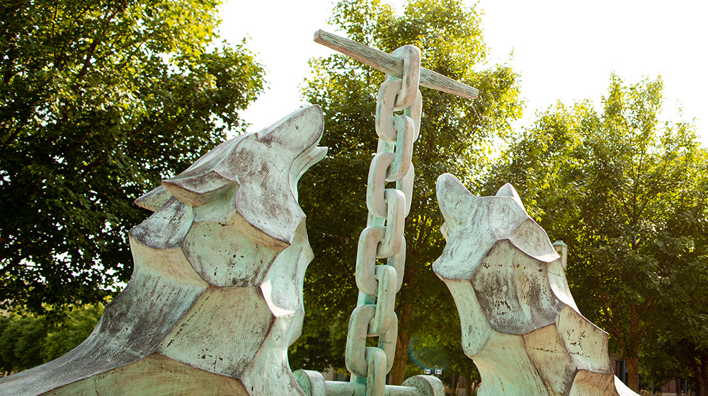 Close-up of the Los Lobos statue depicting the Loyola family crest of the wolf and kettle against a backdrop of trees on Lake Shore Campus.