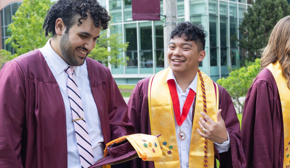 Two students laugh in their graduation regalia and Rambler Journey pins and stoles on Lake Shore Campus. Two students laugh in their graduation regalia and Rambler Journey pins and stoles on Lake Shore Campus.
