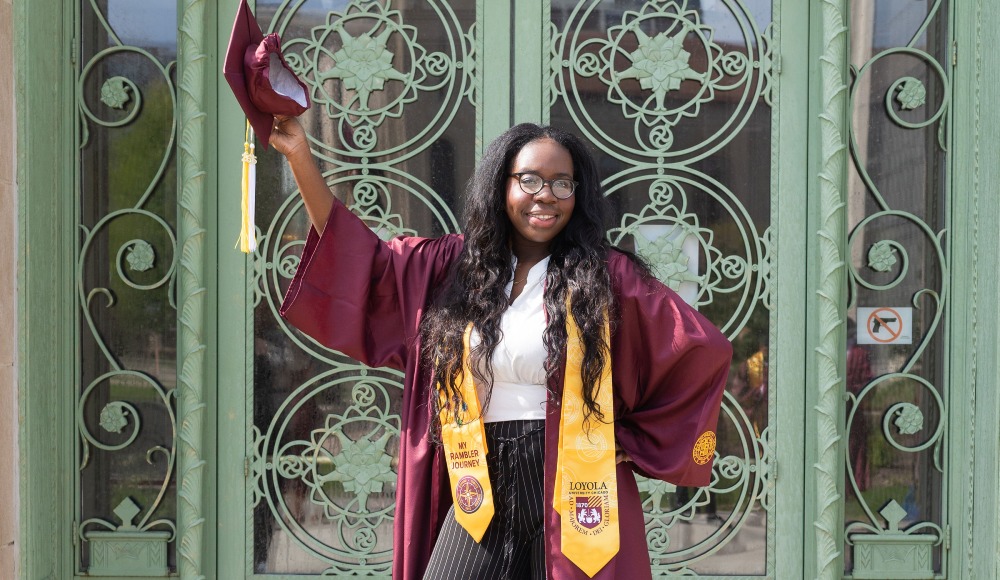 A student wearing graduation regalia holds her cap over her head outside of the green doors of Cudahy Library. A student wearing graduation regalia holds her cap over her head outside of the green doors of Cudahy Library.