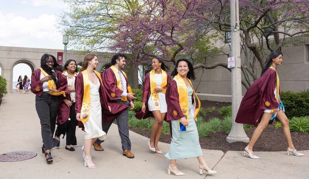 Students wearing graduation regalia and the Rambler Journey pins and stoles walk outside of Madonna Della Strada chapel.