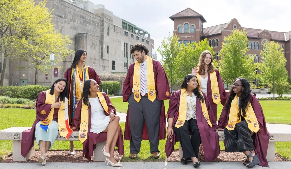 Students wearing graduation regalia and the Rambler Journey pins and stoles sit on benches in a grassy area on Lake Shore Campus.