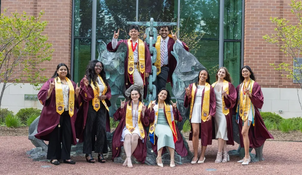 Students wear the Rambler Journey stoles and pins over their graduation regalia as they pose for a photo at the Wolf and Kettle statue.