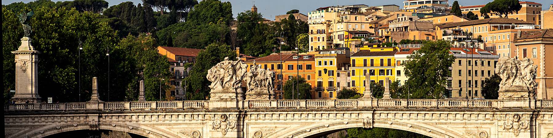 A village in Rome sits in the background behind an ornate bridge that crosses a river just outside of the village.