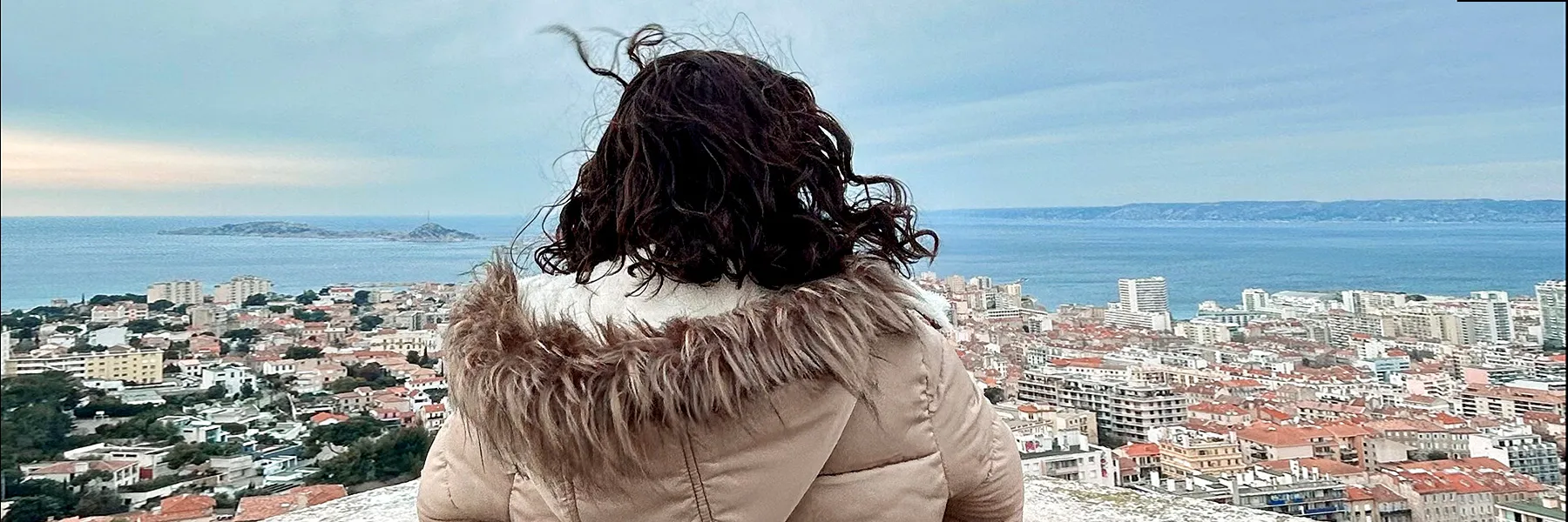 A student in a tan coat stands on an overlook gazing outward to a village that lines the ocean shore.