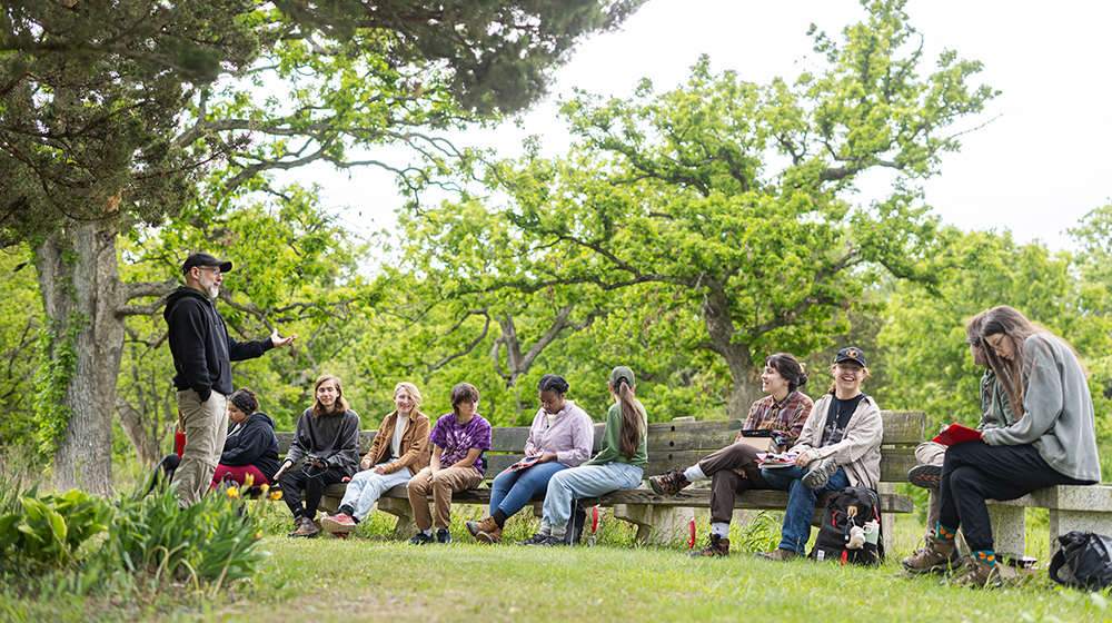 أستاذ يتحدث إلى مجموعة من الطلاب الجالسين على مقاعد في الهواء الطلق