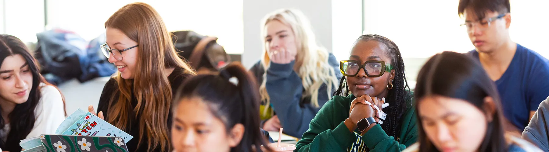 A very attentive young African American woman sits in a group of several students. She is wearing a dark green sweatshirt. All of the students sit at tables.