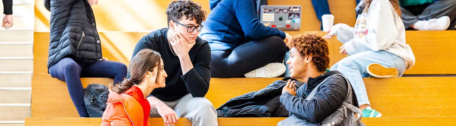 Several students sit on the benches in the Quinlan School of Business atrium. In the foreground there is a young woman and two young men conversing. The woman wears an orange sweater, The young man in the middle has dark curly hair and he wears a dark pullover shirt. The young man on the right has curly brown hair and wear a light weight coat.