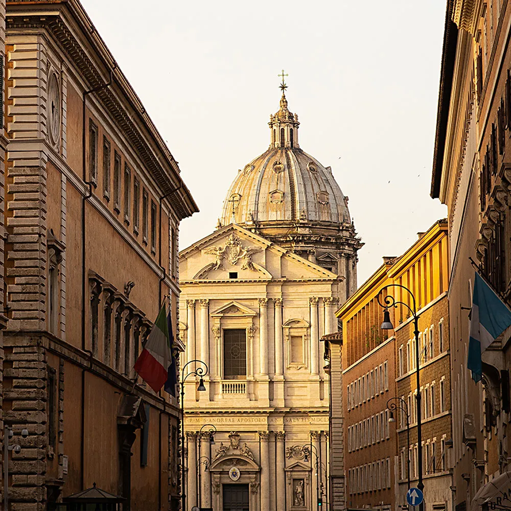 Улица в Риме, вымощенная историческими зданиями, с куполообразным зданием на дальнем расстоянии.