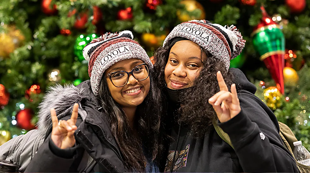 Two young, African American women, lean closely toward one another as they make the wolf sign posing in front of the colorful, decorated tree.