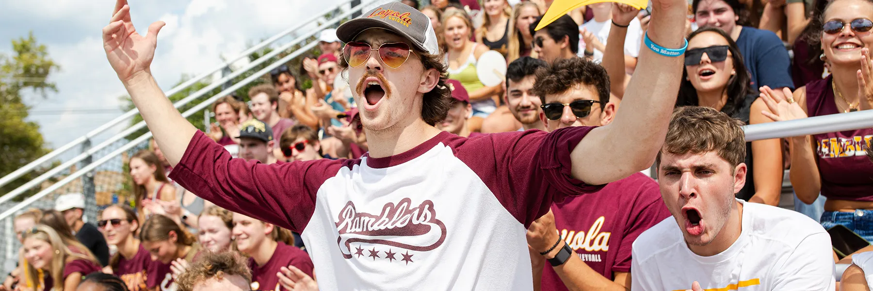 A student in a maroon and white Loyola baseball shirt, wearing a Loyola baseball cap shouts at the Hustle to Hoyne event during Welcome Week.