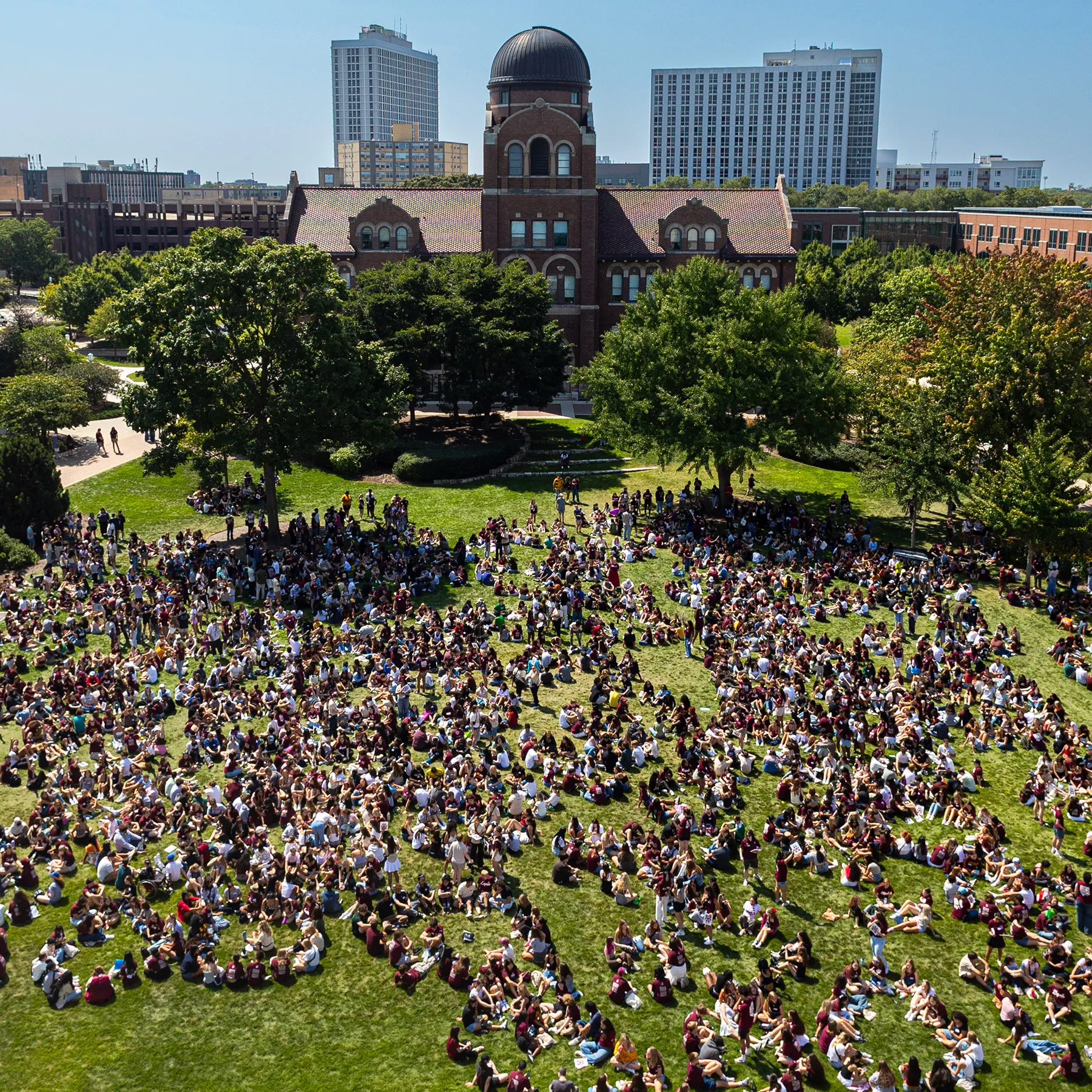 An aerial perspective of hundreds of new Loyola students standing and sitting in groups on the lawn assemble across the open lawn of the Lake Shore Campus. Campus buildings encircle the lawn. Lake Michigan fills the background