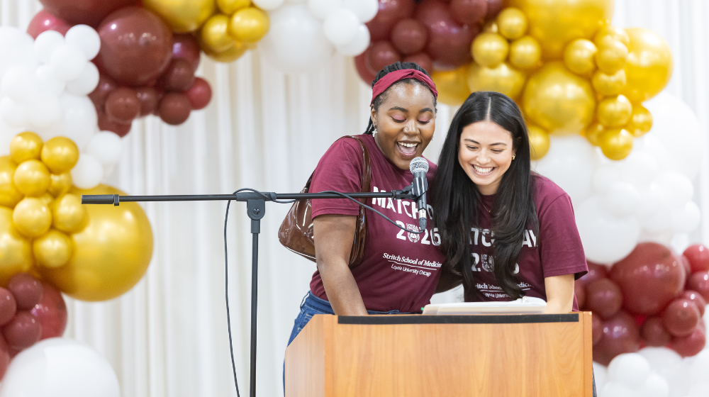 M4 Class President Athena Barrett (right) wears a maroon shirt and stands at a podium alongside M4 Class Vice President Phaedra Johnson (left).