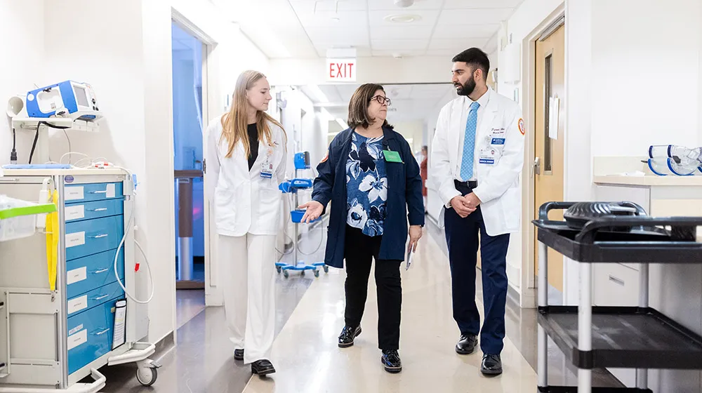 Two medical students wearing white coats walking down a hospital hallway with a Chaplain wearing a blue coat in the middle of the two students