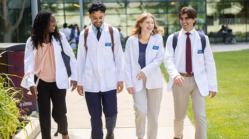 Four medical students wearing white coats walking and talking on campus. Four medical students wearing white coats walking and talking on campus.
