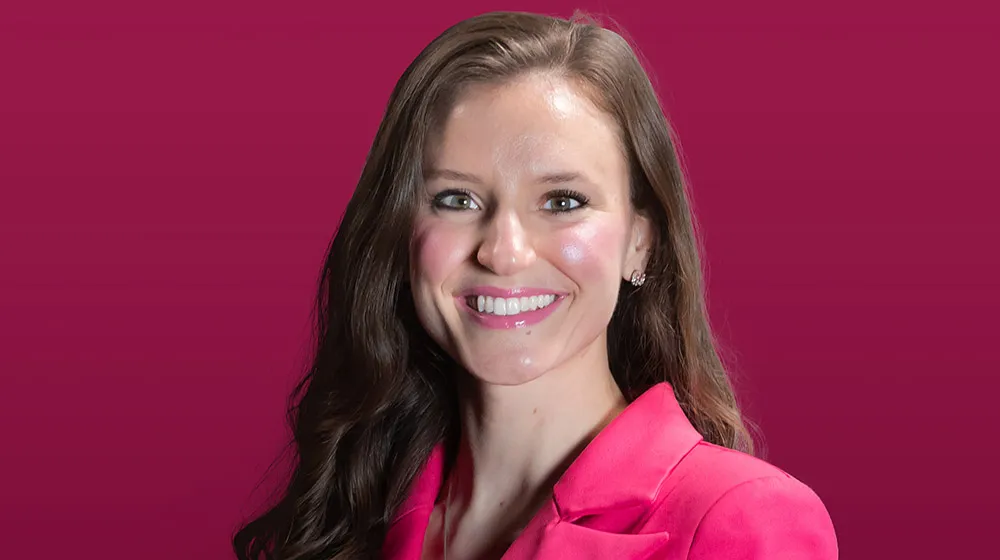 Megi Maci wearing a magenta suit smiling in front of a maroon background