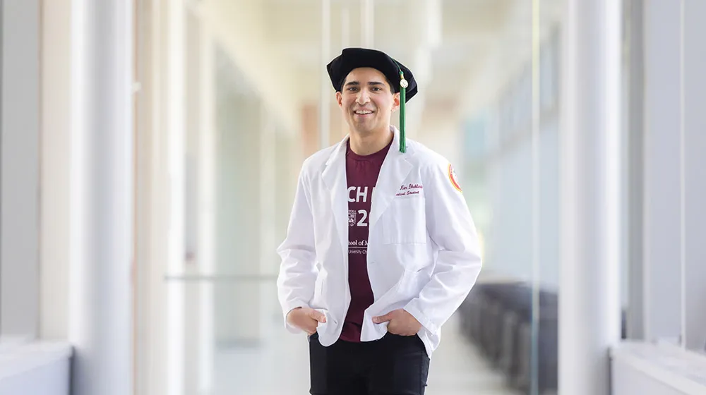 Dual MD/MPH student Nathan Dhablania standing in a hallway wearing a maroon Match Day t-shirt, his white medical coat, and his graduation cap