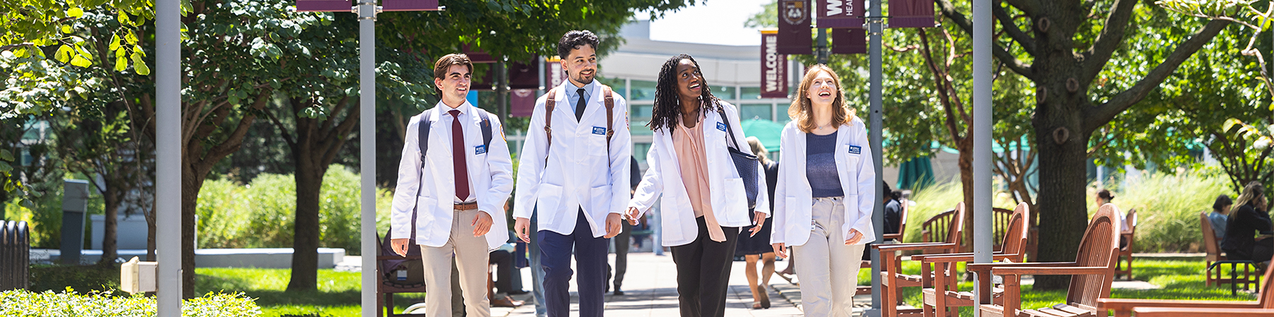 Four medical students, dressed in white lab coats, talk as they walk through campus.