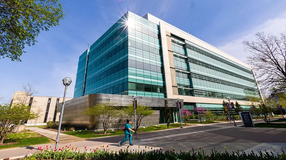 A contemporary glass campus building with trees and flowers, as a person walks along the sidewalk