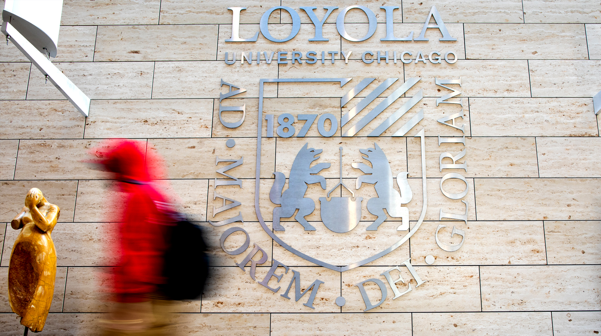 A person in motion walks past a large Loyola University Chicago wall emblem featuring the school’s crest, motto, and founding year.