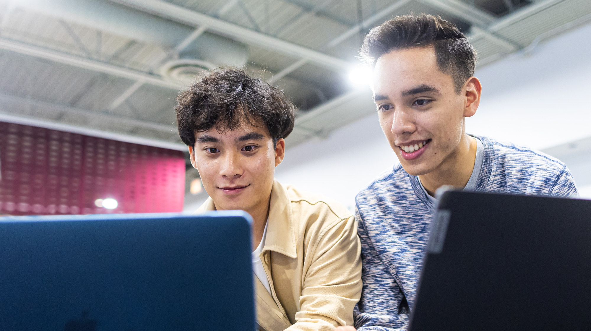 Two students sit closely together working on laptops in a bright indoor space, focusing on their screens beneath overhead industrial-style lighting.