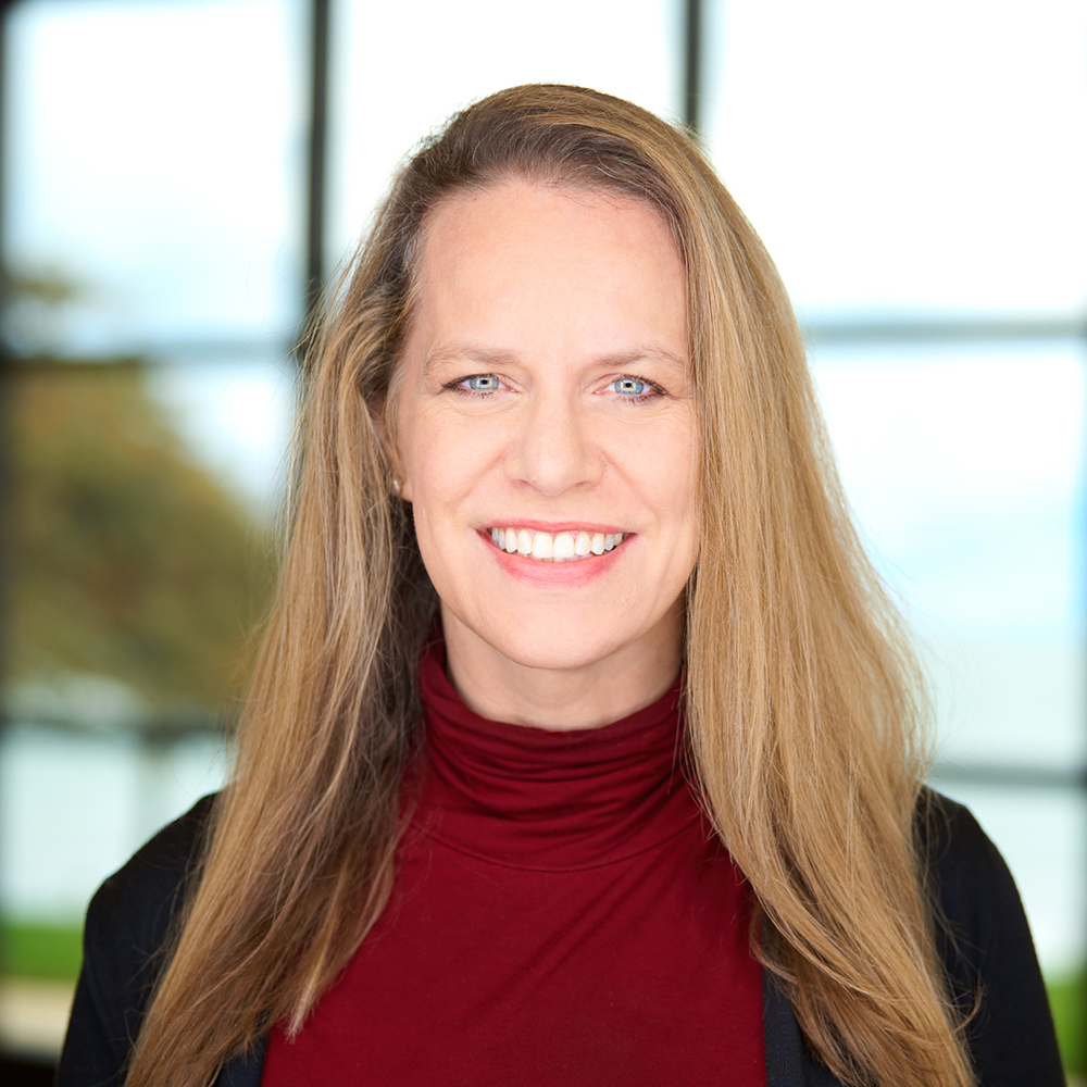 Professional headshot of a smiling woman with long blonde hair wearing a red top against a softly blurred outdoor background