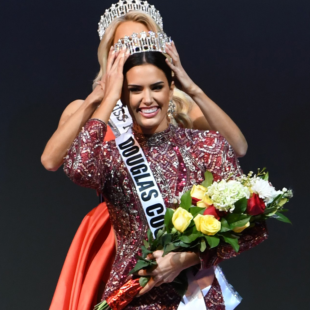 Woman crowned at pageant, wearing sash and holding bouquet of roses on stage