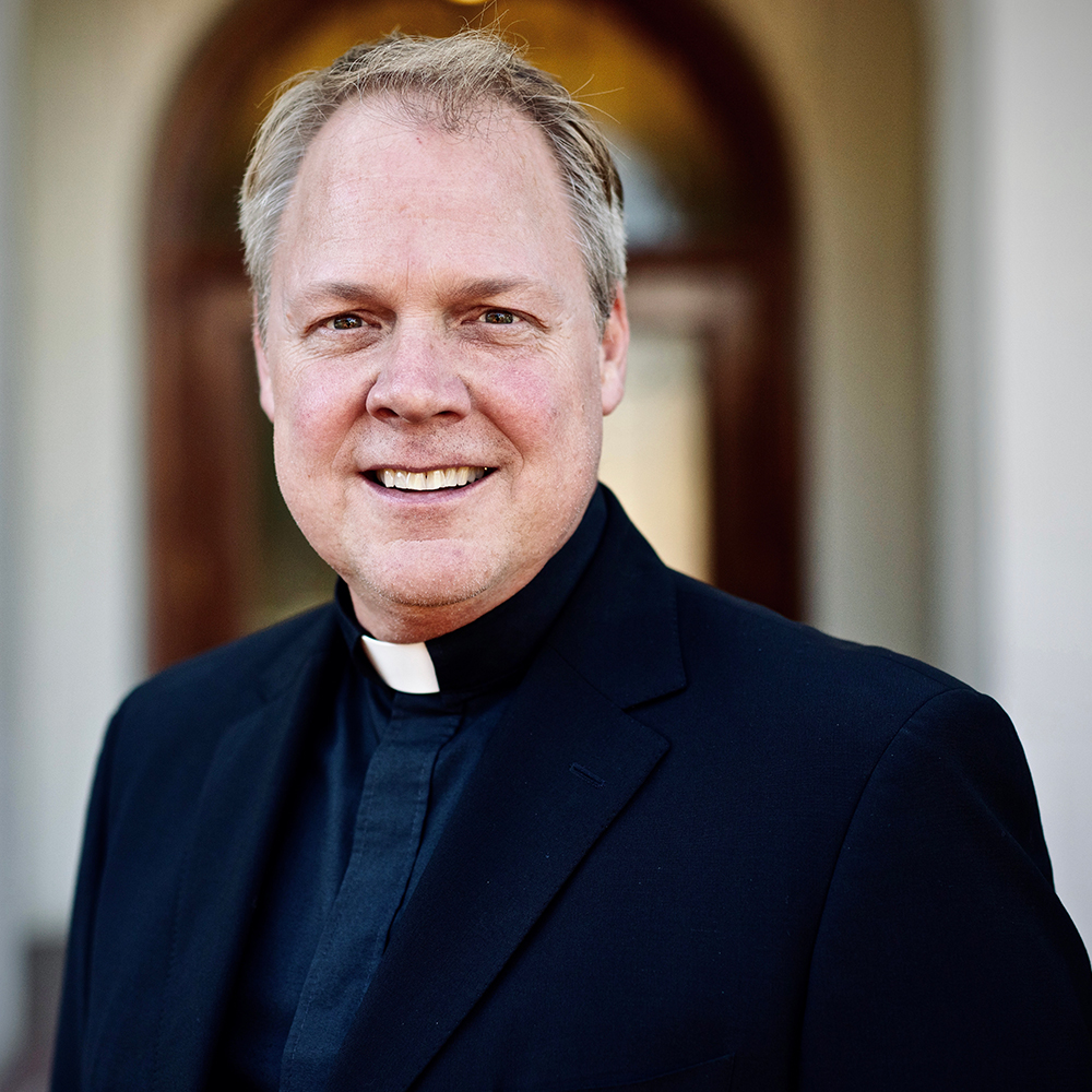 Professional headshot of a smiling Catholic priest in a black clerical collar, standing outdoors in front of an arched building entrance