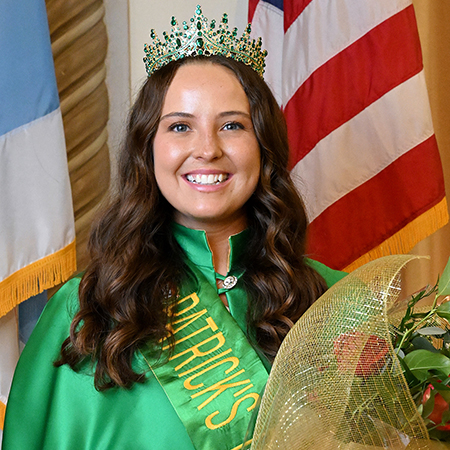 Woman wearing a jeweled crown partially obscured by flowers, with a flag in the background