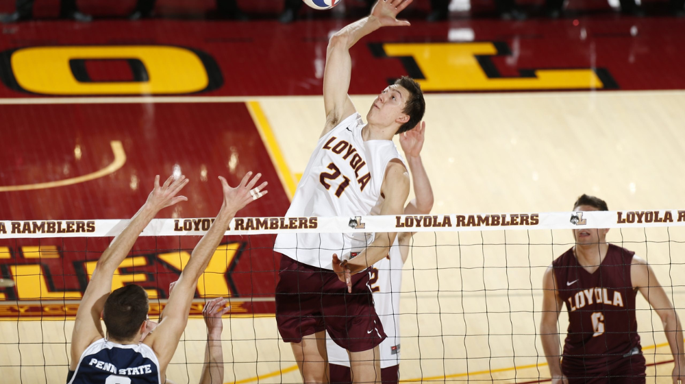 Loyola men’s volleyball player jumps to spike the ball over the net during a match, with opposing blockers attempting to defend
