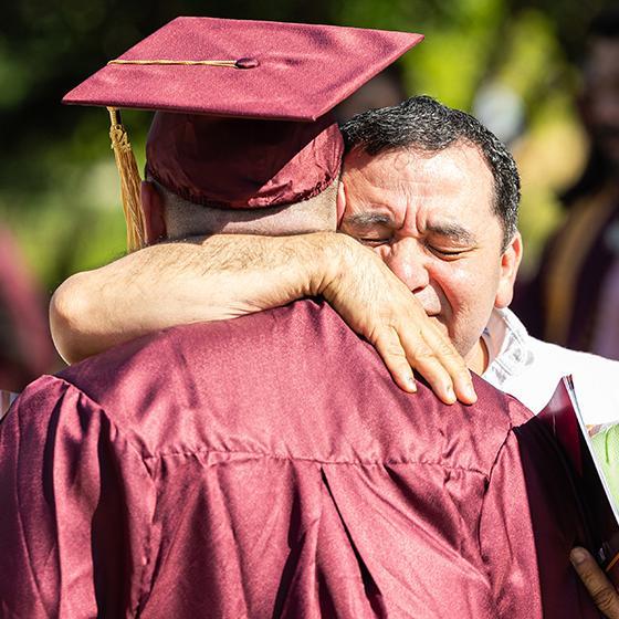 Graduate in cap and gown embraces an emotional family member outdoors at commencement Graduate in cap and gown embraces an emotional family member outdoors at commencement